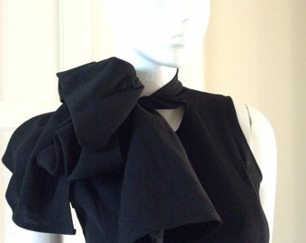 May include: A black sleeveless dress with a large, dramatic bow at the neckline. The bow is made of the same fabric as the dress and is tied in a loose knot.
