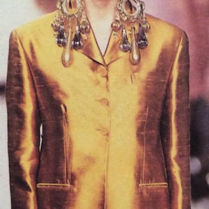 Può includere: Una donna indossa un blazer di raso dorato con un davanti lungo e aperto. Indossa grandi orecchini pendenti con pietre dorate e trasparenti.