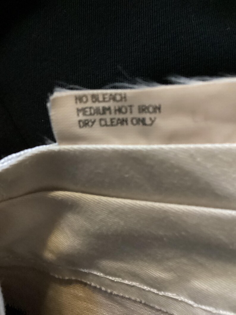 Puede incluir: Primer plano de una etiqueta de cuidado de la tela blanca con el texto "NO BLEACH MEDIUM HOT IRON DRY CLEAN ONLY".