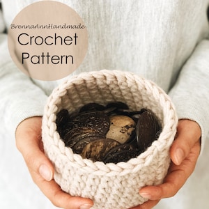 May include: A close-up of a crocheted basket in a light beige color, held by hands. The basket is filled with small, round, brown objects.