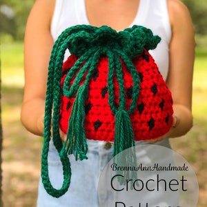 Könnte beinhalten: Eine gehäkelte Tasche in Form einer Erdbeere mit grünem Kordelzug und Quaste. Die Tasche wird von einer Person gehalten, die ein weißes Tanktop und eine blaue Jeans trägt. Der Text "BrennaAnnHandmade Crochet Pattern" ist im Bild sichtbar.
