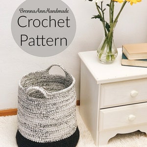 Puede incluir: Una cesta de almacenamiento de crochet con dos asas, con un esquema de color blanco y negro. La cesta está sentada en una alfombra blanca junto a una mesita de noche blanca con dos cajones. Un jarrón de flores amarillas y blancas se sienta en la mesita de noche.