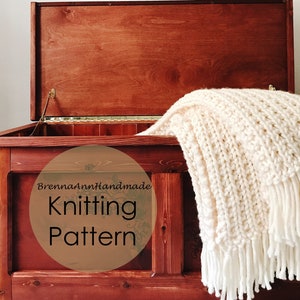 May include: A wooden storage chest with a white knitted blanket inside. The chest has a hinged lid and is made of dark wood. The blanket has a textured pattern and is fringed. The text "BrennaAnnHandmade Knitting Pattern" is on a circle in the front of the chest.