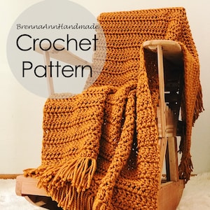 May include: A crocheted throw blanket in a mustard yellow colour with fringe on the edges. The blanket is draped over a wooden rocking chair. The text "BrennaAnnHandmade Crochet Pattern" is visible in the image.
