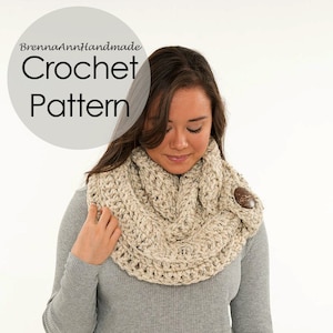 Op de afbeelding: Een beige oneindige sjaal van gehaakt garen met een houten knoop. De sjaal wordt gedragen door een persoon met lang bruin haar. De tekst "BrennaAnnHandmade Crochet Pattern" staat in een cirkel boven het hoofd van de persoon.