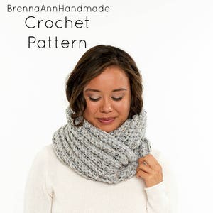 May include: A grey and white crocheted infinity scarf worn by a person. The scarf is thick and textured. The text "BrennaAnnHandmade Crochet Pattern" is at the top of the image.