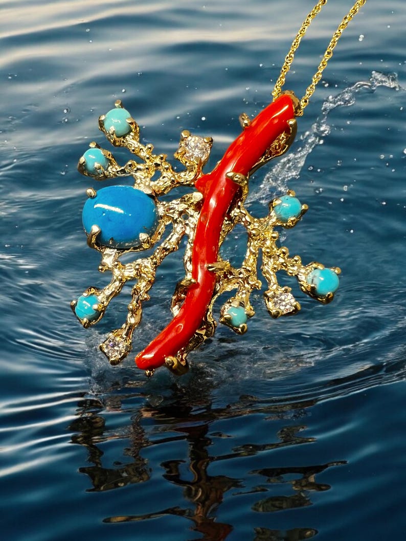 May include: A gold pendant necklace with a red coral branch and turquoise gemstones. The pendant is shaped like a tree branch with a red coral branch extending from the center. The pendant is hanging from a gold chain and is suspended in the air above a blue water surface.