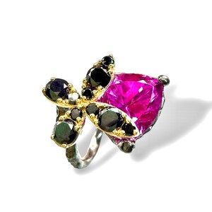 Puede incluir: Un anillo de plata con una gran gema fucsia en forma de coraz&oacute;n. El anillo presenta un dise&ntilde;o floral de gemas negras y metal dorado, creando un contraste llamativo. El anillo est&aacute; sobre un fondo blanco.