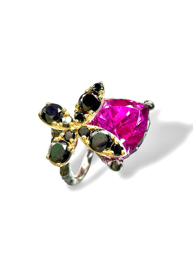 Puede incluir: Un anillo de plata con una gran gema fucsia en forma de coraz&oacute;n. El anillo presenta un dise&ntilde;o floral de gemas negras y metal dorado, creando un contraste llamativo. El anillo est&aacute; sobre un fondo blanco.