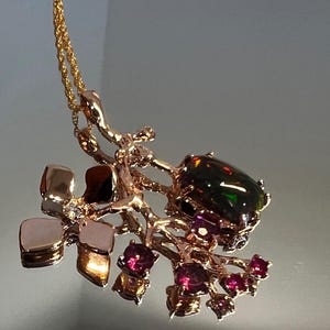 May include: A gold-coloured pendant necklace with a dark, iridescent gemstone and smaller red gemstones. The pendant has an organic, branch-like design with a gold chain. The necklace is a piece of jewellery.