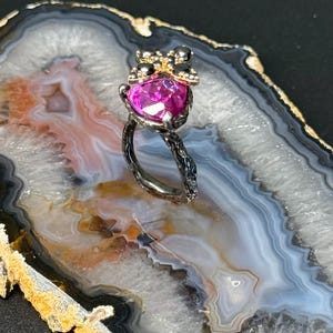 Puede incluir: Un anillo plateado con una gran piedra preciosa fucsia en forma de coraz&oacute;n. El engaste del anillo presenta un dise&ntilde;o floral en la banda y un grupo de peque&ntilde;as piedras negras y detalles dorados sobre la piedra preciosa. El anillo se exhibe sobre una superficie veteada.