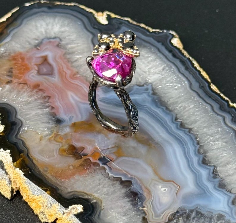 Puede incluir: Un anillo plateado con una gran piedra preciosa fucsia en forma de coraz&oacute;n. El engaste del anillo presenta un dise&ntilde;o floral en la banda y un grupo de peque&ntilde;as piedras negras y detalles dorados sobre la piedra preciosa. El anillo se exhibe sobre una superficie veteada.