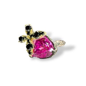 Puede incluir: Primer plano de un anillo con una gran gema rosa facetada. El anillo tiene un engaste dorado con gemas negras y una banda plateada. El anillo est&aacute; sobre un fondo blanco.