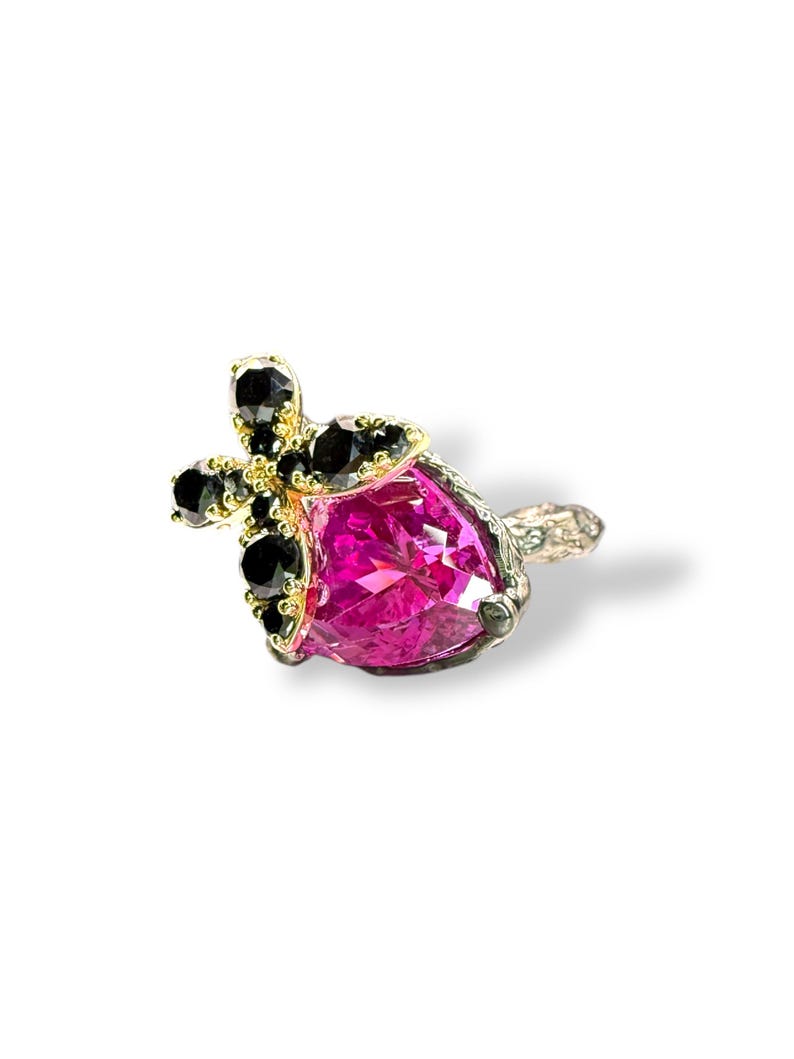 Puede incluir: Primer plano de un anillo con una gran gema rosa facetada. El anillo tiene un engaste dorado con gemas negras y una banda plateada. El anillo est&aacute; sobre un fondo blanco.