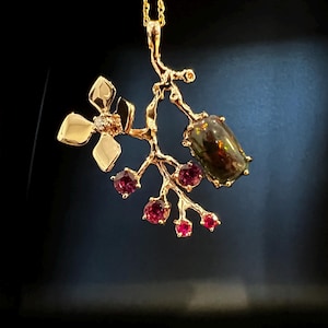 May include: A gold pendant necklace with a botanical design. The pendant features a dark gemstone, small red gemstones, and a flower with a diamond. The necklace is suspended from a fine gold chain. The pendant's design resembles a branch.
