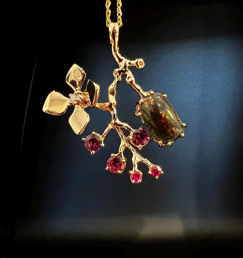 May include: A gold pendant necklace with a botanical design. The pendant features a dark gemstone, small red gemstones, and a flower with a diamond. The necklace is suspended from a fine gold chain. The pendant's design resembles a branch.