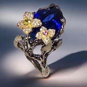 Puede incluir: Un anillo de plata con una gran piedra preciosa azul oscuro. Dos pequeños acentos florales dorados con diminutas piedras preciosas están engastados en el lateral de la piedra azul. El anillo tiene un diseño orgánico texturizado.