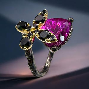 Puede incluir: Un anillo de plata con una gran piedra preciosa magenta en forma de coraz&oacute;n. El anillo est&aacute; adornado con un grupo de piedras preciosas negras y metal dorado, creando un dise&ntilde;o floral. La banda del anillo tiene una apariencia texturizada y org&aacute;nica.