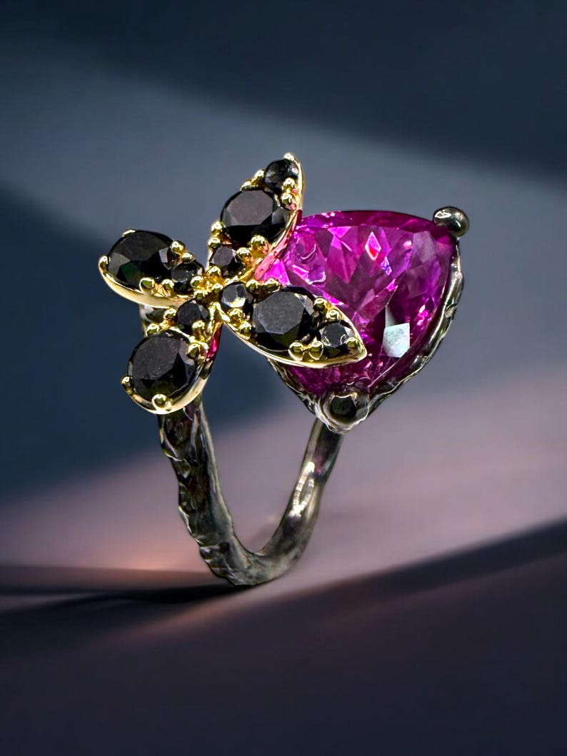 Puede incluir: Un anillo de plata con una gran piedra preciosa magenta en forma de coraz&oacute;n. El anillo est&aacute; adornado con un grupo de piedras preciosas negras y metal dorado, creando un dise&ntilde;o floral. La banda del anillo tiene una apariencia texturizada y org&aacute;nica.