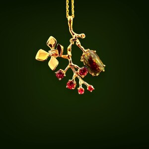 May include: A gold pendant necklace with a floral and berry motif. The pendant features a flower with a small gemstone, a branch with red gemstones, and a larger, dark-coloured gemstone. The necklace hangs on a delicate gold chain.