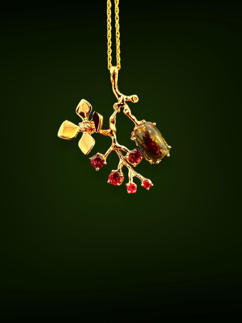 May include: A gold pendant necklace with a floral and berry motif. The pendant features a flower with a small gemstone, a branch with red gemstones, and a larger, dark-coloured gemstone. The necklace hangs on a delicate gold chain.