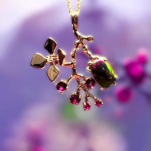 May include: A gold pendant necklace with a floral design. The pendant features a branch-like structure with gold leaves, small pink gemstones, and a larger, dark green and black gemstone. The necklace hangs on a delicate gold chain.