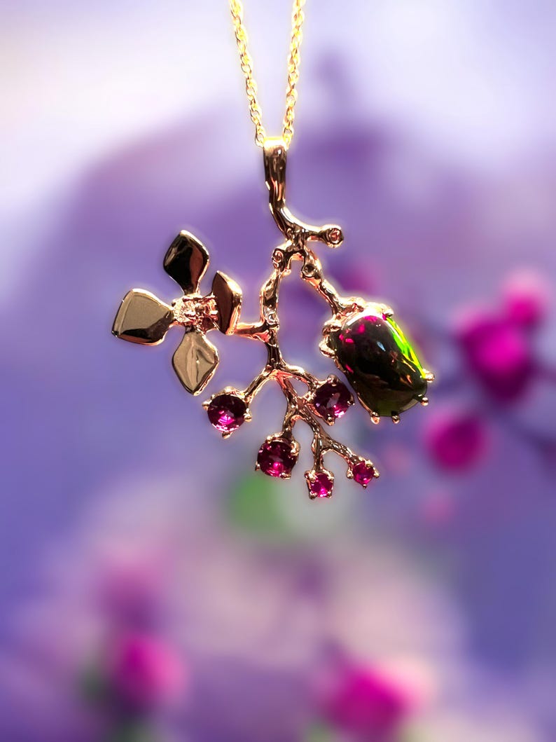 May include: A gold pendant necklace with a floral design. The pendant features a branch-like structure with gold leaves, small pink gemstones, and a larger, dark green and black gemstone. The necklace hangs on a delicate gold chain.
