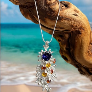 May include: A silver pendant necklace featuring a blue gemstone and smaller yellow gemstones set in a coral-like design. The necklace is suspended from a silver chain.