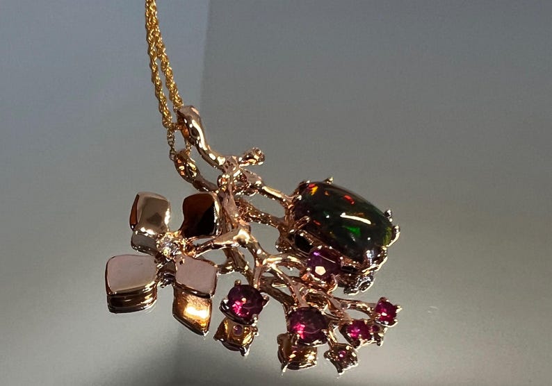 May include: A gold-coloured pendant necklace with a dark, iridescent gemstone and smaller red gemstones. The pendant has an organic, branch-like design with a gold chain. The necklace is a piece of jewellery.