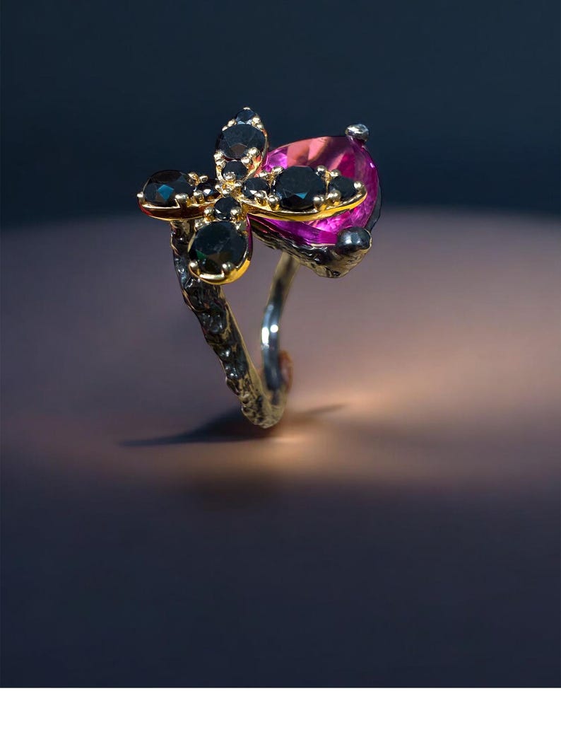 Puede incluir: Un anillo &uacute;nico con un dise&ntilde;o floral dorado con piedras negras y una gran piedra rosa en forma de l&aacute;grima. La banda del anillo es plateada y el engaste tiene una apariencia texturizada y org&aacute;nica.