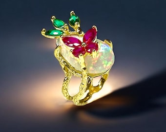 May include: A gold ring with an oval, iridescent gemstone. The ring is decorated with a butterfly made of red gemstones and green leaf accents. The ring's band has a textured, organic design. The ring is a piece of jewellery.