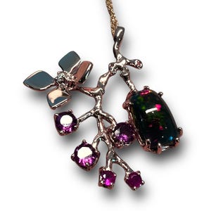 May include: A silver pendant necklace with a floral design. It features a large, dark opal gemstone and smaller purple gemstones. The necklace has a delicate chain and is set against a white background.