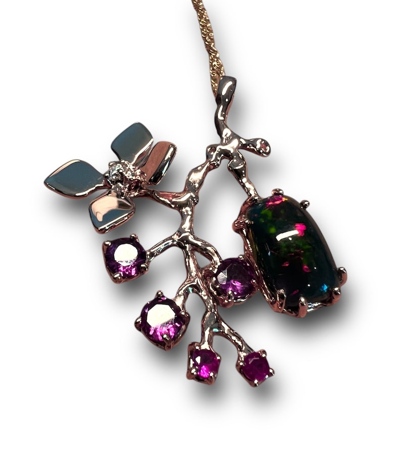 May include: A silver pendant necklace with a floral design. It features a large, dark opal gemstone and smaller purple gemstones. The necklace has a delicate chain and is set against a white background.
