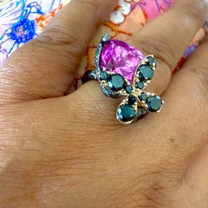 Puede incluir: Un anillo plateado con una gran piedra preciosa magenta en forma de pera. El anillo est&aacute; adornado con un dise&ntilde;o de mariposa, con piedras preciosas de color verde azulado oscuro que forman las alas. El anillo est&aacute; en una mano.