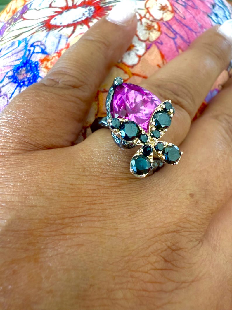 Puede incluir: Un anillo plateado con una gran piedra preciosa magenta en forma de pera. El anillo est&aacute; adornado con un dise&ntilde;o de mariposa, con piedras preciosas de color verde azulado oscuro que forman las alas. El anillo est&aacute; en una mano.