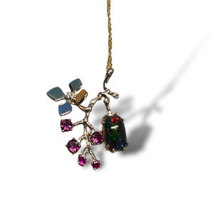 May include: A gold pendant necklace with a branch design, a large dark opal gemstone, and small pink gemstones. The pendant also features a flower with blue and brown accents. The necklace is suspended from a fine gold chain.