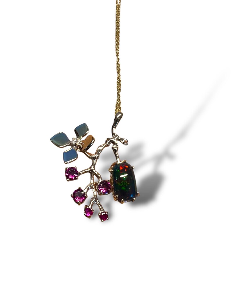 May include: A gold pendant necklace with a branch design, a large dark opal gemstone, and small pink gemstones. The pendant also features a flower with blue and brown accents. The necklace is suspended from a fine gold chain.