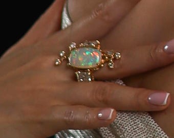 May include: A gold ring featuring a large, iridescent opal gemstone surrounded by smaller diamonds. The ring is worn on a finger with a white manicure.
