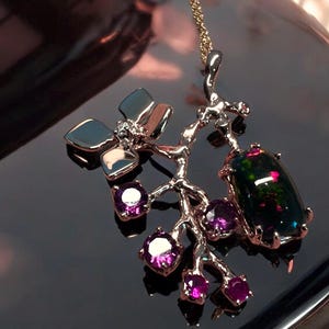 May include: A gold-coloured pendant necklace. It features a branch design with a large, dark opal gemstone and smaller purple gemstones. The necklace also includes silver-coloured leaf-shaped accents and a delicate gold chain.