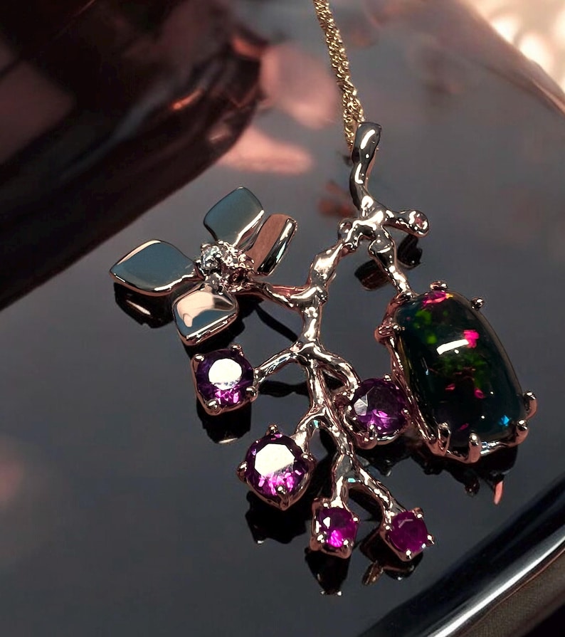 May include: A gold-coloured pendant necklace. It features a branch design with a large, dark opal gemstone and smaller purple gemstones. The necklace also includes silver-coloured leaf-shaped accents and a delicate gold chain.