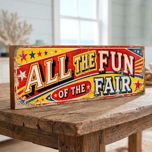 May include: A rectangular wooden sign with a distressed finish featuring the text "ALL THE FUN OF THE FAIR" in bold, multi-coloured lettering. The design includes red, yellow, and blue accents with star graphics against a weathered background.