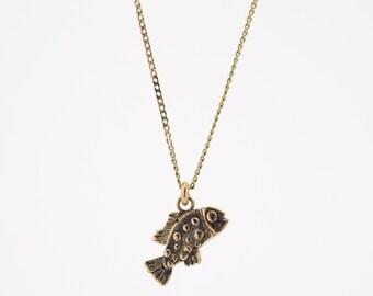 Könnte beinhalten: Eine goldene Kettenkette mit einem kleinen, detaillierten Fisch-Anhänger. Der Fisch ist strukturiert und hat eine leicht oxidierte Oberfläche.