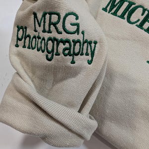 May include: A beige sweatshirt with the embroidered text "MRG Photography" in green on the sleeve. The word "MICHAEL" is also embroidered in green on the garment. The fabric is folded, showing the texture of the material.