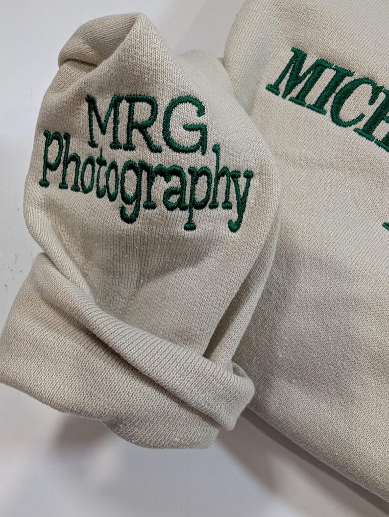 May include: A beige sweatshirt with the embroidered text "MRG Photography" in green on the sleeve. The word "MICHAEL" is also embroidered in green on the garment. The fabric is folded, showing the texture of the material.