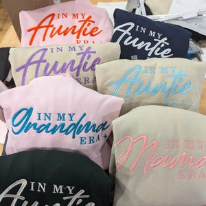 May include: A collection of seven sweatshirts with embroidered text. The sweatshirts are in various colors, including pink, beige, navy blue, and black. The text on the sweatshirts reads "In My Auntie Era", "In My Grandma Era", and "In My Mama Era".
