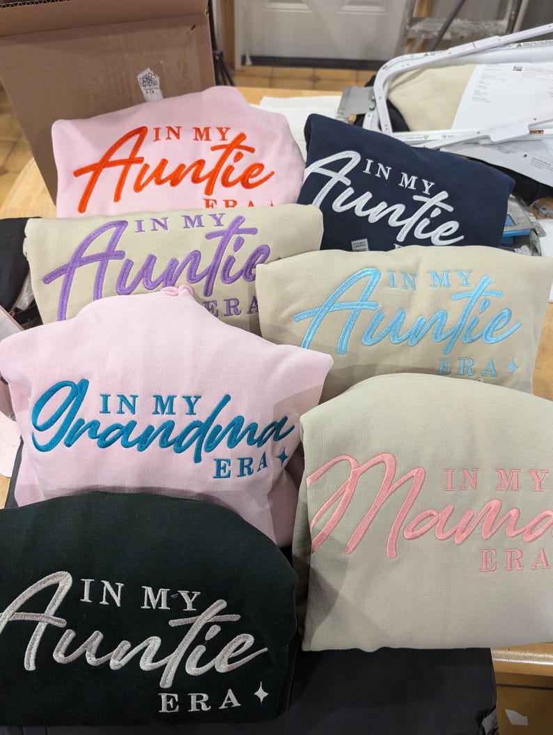 May include: A collection of seven sweatshirts with embroidered text. The sweatshirts are in various colors, including pink, beige, navy blue, and black. The text on the sweatshirts reads "In My Auntie Era", "In My Grandma Era", and "In My Mama Era".