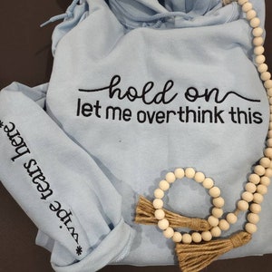 May include: Light blue hoodie with the text "hold on let me overthink this" embroidered on the front. The hoodie has a drawstring hood and a small embroidered design on the sleeve that says "wipe tears here". A wooden beaded necklace with tassels is draped over the hoodie.