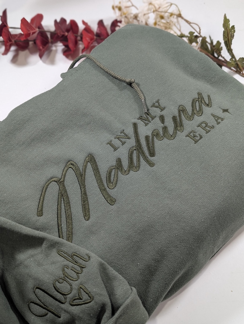 May include: A green hooded sweatshirt with the text "In My Madrina Era" embroidered on the front and "Noah" embroidered on the left sleeve.