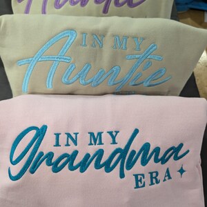 May include: Three sweatshirts with embroidered text. The top sweatshirt is purple with the text "In My Auntie Era". The middle sweatshirt is beige with the text "In My Auntie Era". The bottom sweatshirt is pink with the text "In My Grandma Era".