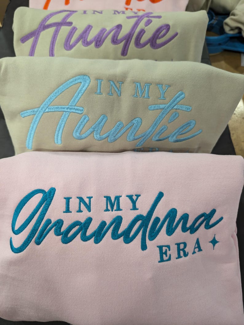 May include: Three sweatshirts with embroidered text. The top sweatshirt is purple with the text "In My Auntie Era". The middle sweatshirt is beige with the text "In My Auntie Era". The bottom sweatshirt is pink with the text "In My Grandma Era".